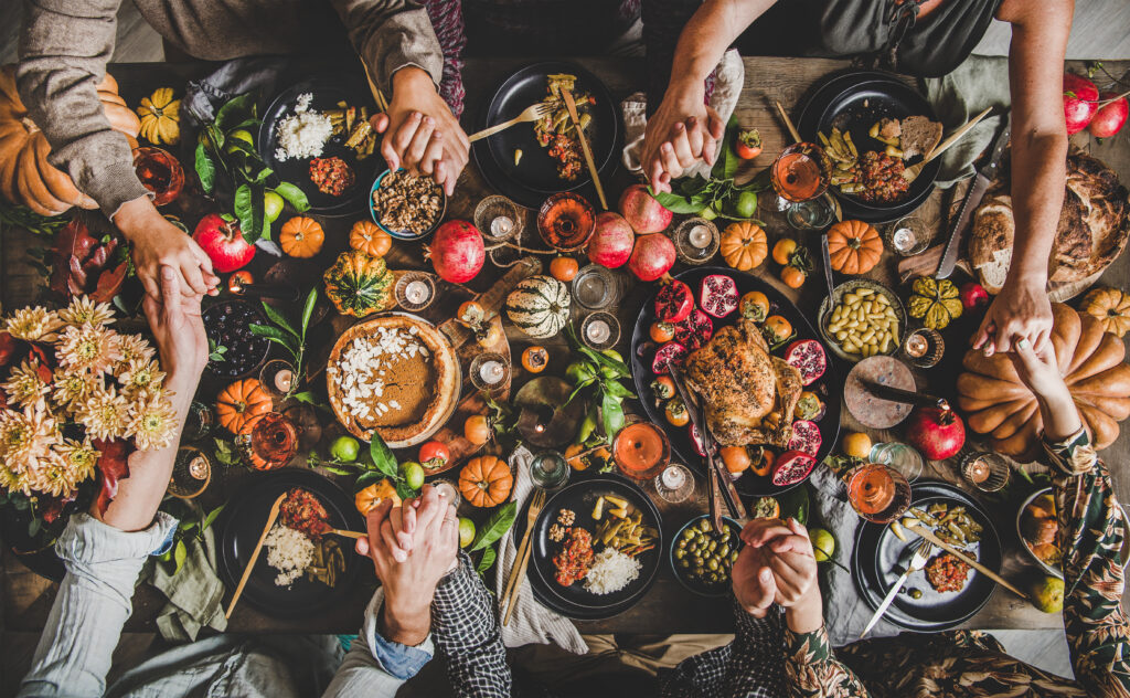 Family praying holding hands at Thanksgiving table. Flat-lay of feasting peoples hands over Friendsgiving table with Autumn food, candles, roasted turkey and pumpkin pie over wooden table, top view